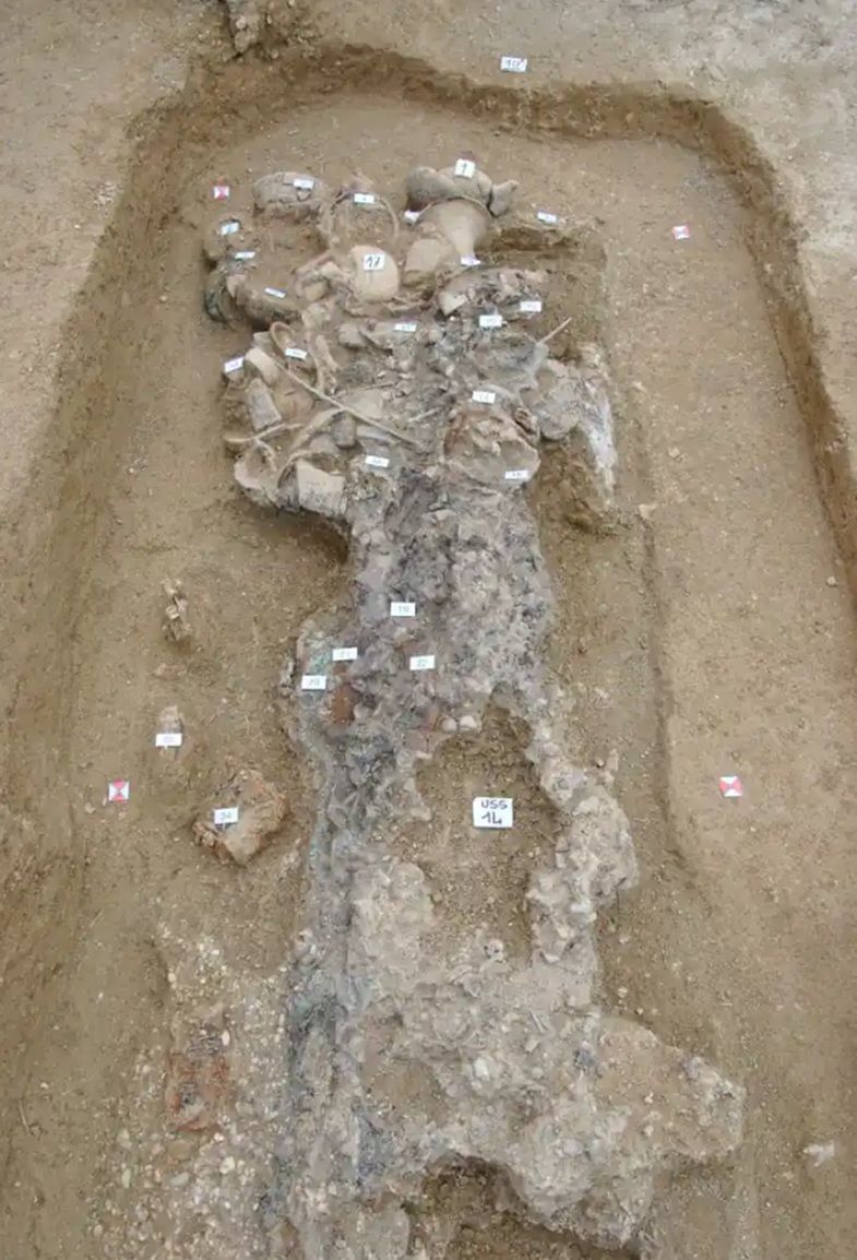 In einer rechteckigen Erdgrube liegt ein menschliches Skelett, das von zahlreichen Keramikgefäßen und Grabbeigaben umgeben ist. Die Knochen sind teilweise von Erde bedeckt und zeigen Verfärbungen. Mehrere kleine, nummerierte Markierungen sind auf den Fundstücken und im Bereich des Skeletts angebracht.