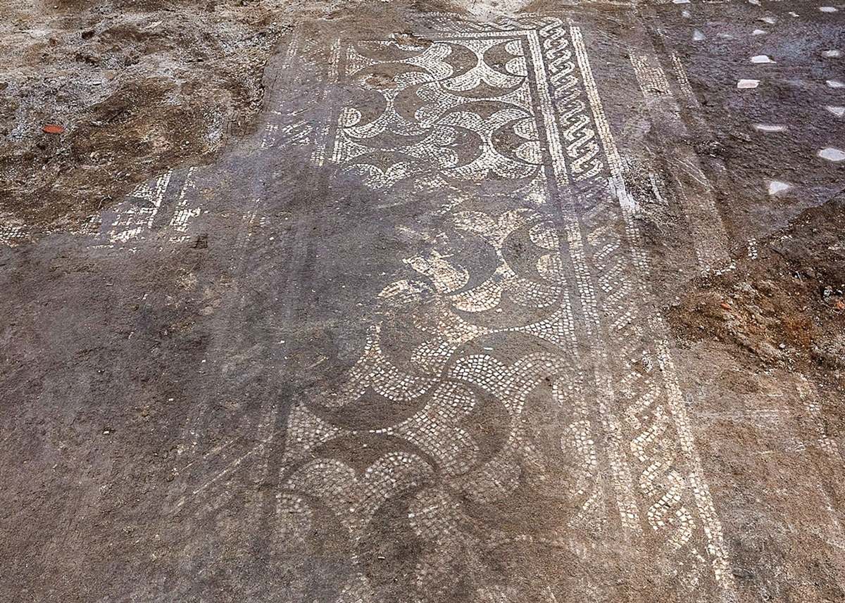 Ein rechteckiges Bodenmosaik mit geometrischen und wellenförmigen Mustern ist freigelegt. Die Mosaiksteine sind hell und kontrastieren mit dem dunkleren Erdreich. Das Muster besteht aus ineinander verschlungenen Linien und Flächen, die sich über die gesamte Fläche erstrecken. Am Rand sind weitere dekorative Elemente sichtbar.