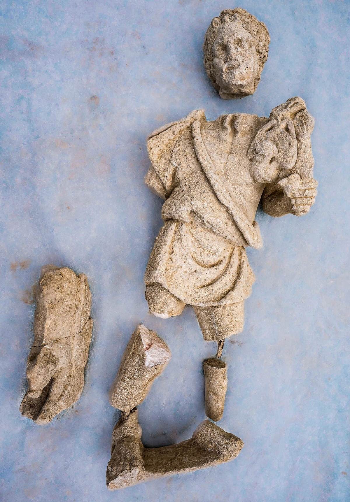 Fragmentierte antike Statue aus hellem Stein auf hellem Untergrund. Der Oberkörper trägt ein drapiertes Gewand. Kopf und Gliedmaßen sind abgebrochen und liegen teilweise separat daneben. Die Oberfläche zeigt deutliche Abnutzungsspuren.