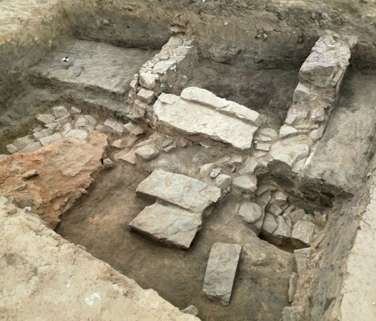 Im Zentrum des Bildes sind freigelegte Mauerreste und Steinplatten zu sehen, die in einem rechteckigen Erdloch liegen. Die Steine sind unterschiedlich groß und teilweise gestapelt, einige wirken wie Fundamente oder Bodenplatten. Der umgebende Boden ist hellbraun und wirkt trocken.