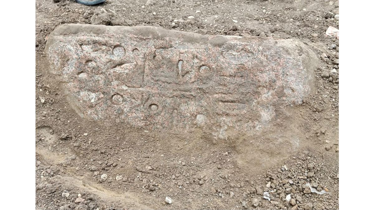 Ein länglicher, teilweise freigelegter Steinblock liegt waagerecht im sandigen Boden. Die Oberfläche zeigt flache Reliefs mit erkennbaren Hieroglyphen und geometrischen Formen.