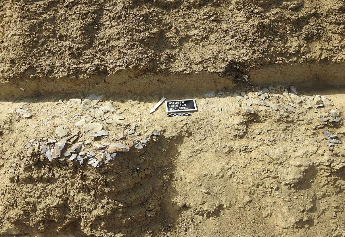 Ein schmaler, horizontal verlaufender Streifen mit zahlreichen kleinen, flachen Steinen und Splittern ist in einer sandfarbenen Erdschicht eingebettet. Oberhalb und unterhalb des Streifens sind kompakte, ungestörte Erdschichten sichtbar. In der Mitte des Streifens liegt ein rechteckiges Schild mit kontrastreichem Text und einer Zahlenreihe.