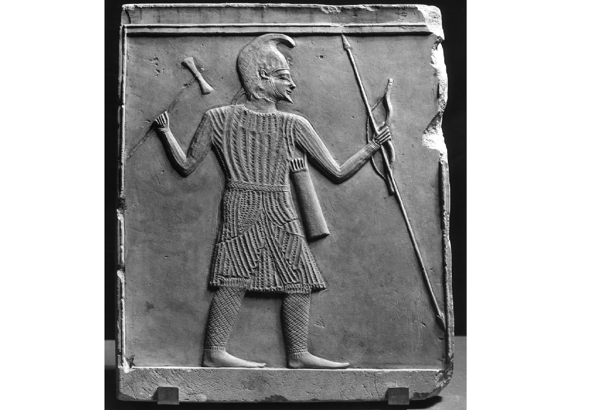 Relief mit einer seitlich dargestellten männlichen Figur in typischer Gewandung. Die Figur trägt ein gemustertes, knielanges Gewand und eine spitz zulaufende Kopfbedeckung. In der linken Hand hält sie einen langen Speer, in der rechten eine Axt. Ein Köcher mit Pfeilen hängt am Rücken. Die Umrisse und Details sind klar herausgearbeitet, der Hintergrund ist glatt. Das Relief ist rechteckig und an den Rändern eingerahmt.