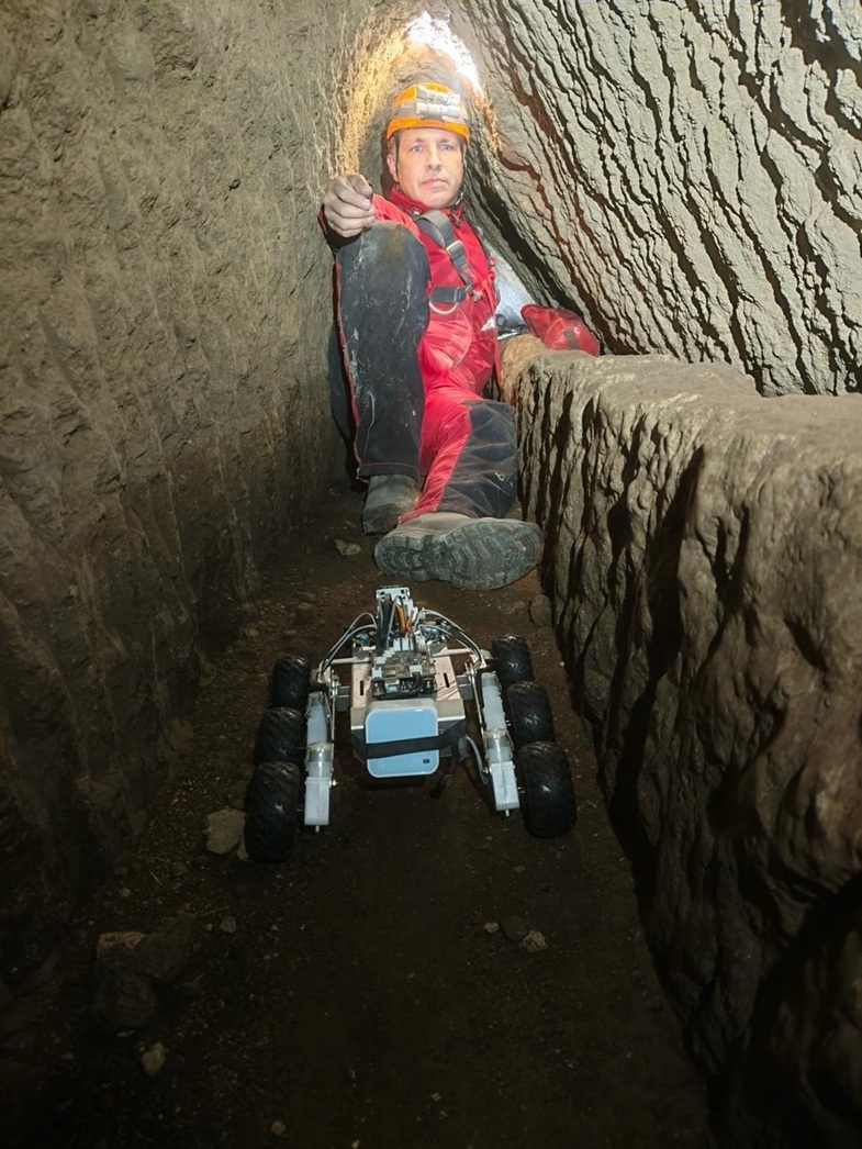 Person in rotem Overall und Helm sitzt in einem engen, steinernen Tunnel hinter einem kleinen, mehrrädrigen Roboterfahrzeug.