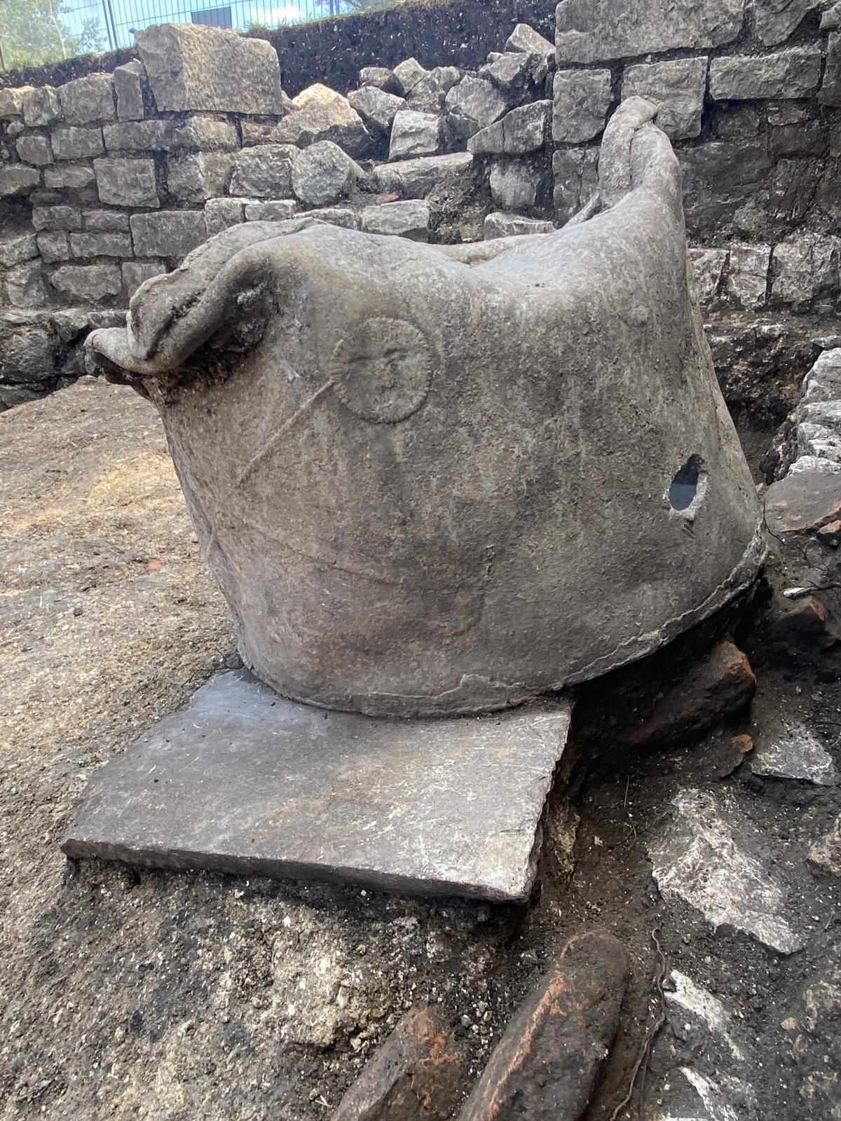 Großer, runder Bleibehälter mit glatter Oberfläche und abgerundeten Kanten, der auf einer rechteckigen Steinplatte steht. In die Vorderseite sind ein Kreis und eine Linie eingeritzt. Links oben befindet sich eine plastische Verzierung, die an einen Tierkopf erinnert. Im Hintergrund sind Mauerreste aus grob behauenen Steinen zu sehen.