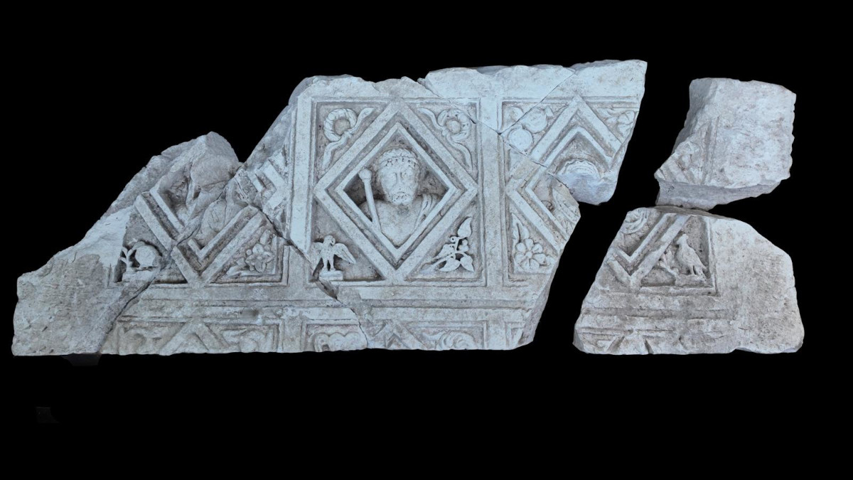 Fragment eines antiken Steinreliefs mit geometrischen Mustern und einem zentralen menschlichen Gesicht in einem Rautenmuster