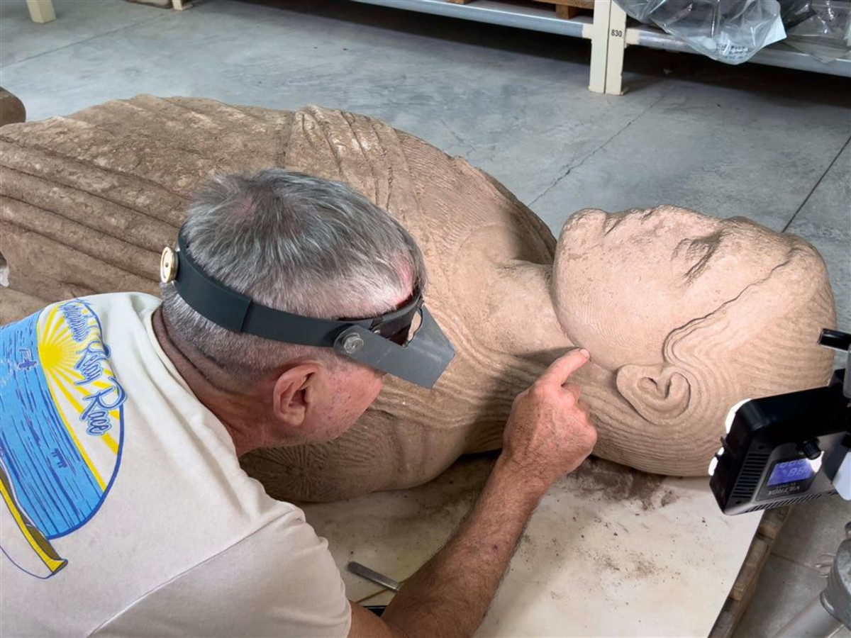 Ein älterer Mann mit grauem Haar und einer Vergrößerungsbrille untersucht eine große, liegende Marmorstatue. Die Statue zeigt eine menschliche Figur mit detailliert ausgearbeitetem Gesicht und gefalteten Gewändern. Der Mann trägt ein helles T-Shirt mit farbigem Aufdruck und berührt mit dem Finger vorsichtig den Halsbereich der Statue. Im Vordergrund steht ein kleines technisches Gerät mit Display, das auf die Statue gerichtet ist. 