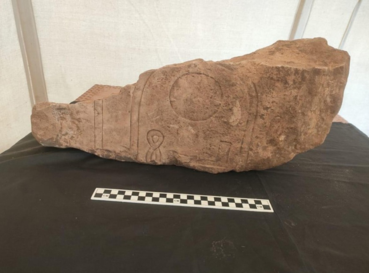 Ein bräunlicher, unregelmäßig geformter Steinblock liegt auf einer dunklen Unterlage. Die Oberfläche zeigt eingravierte Zeichen und Symbole, darunter Kreise und Linien. Vor dem Stein befindet sich eine Messlatte mit abwechselnd schwarzen und weißen Feldern. Im Hintergrund ist eine helle, glatte Fläche sichtbar.