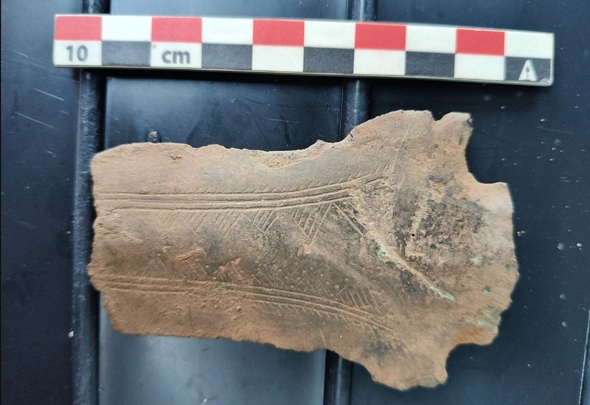 Bruchstück eines braunen Tonscherbens mit eingeritzten Linienmustern, darüber ein Maßstab.