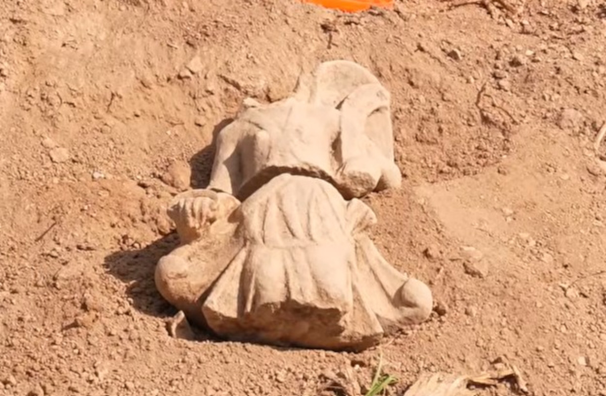 Eine antike Steinskulptur liegt halb freigelegt in sandigem Erdreich. Die Figur zeigt einen Körper mit gefalteter Kleidung und ausgestrecktem Arm, der Kopf fehlt. Die Oberfläche ist hell und weist deutliche Abnutzungsspuren auf.