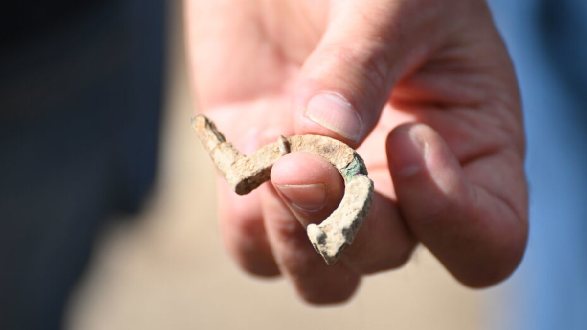 Eine Hand hält ein kleines, gebogenes Fundstück aus hellem, metallenem Material. Die Oberfläche wirkt rau und unregelmäßig, das Objekt ist an mehreren Stellen abgebrochen.