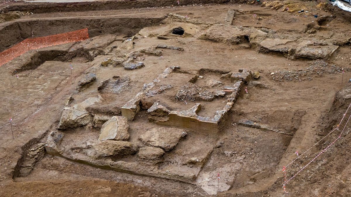 Freigelegte Mauerreste und Fundamente aus hellem Stein liegen in einem großflächig ausgegrabenen Erdreich. Die Strukturen sind unregelmäßig angeordnet und von Gräben und Erdaufschüttungen umgeben.