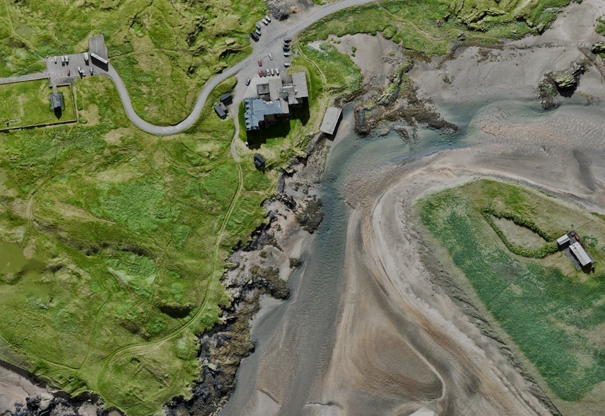 Drohnenaufnahme des Hafenbereichs von Búðir. Zu sehen ist Ein Flussarm mit sandigem Uferbereich, moderne Gebäude mit angrenzendem Parkplatz und Straße.