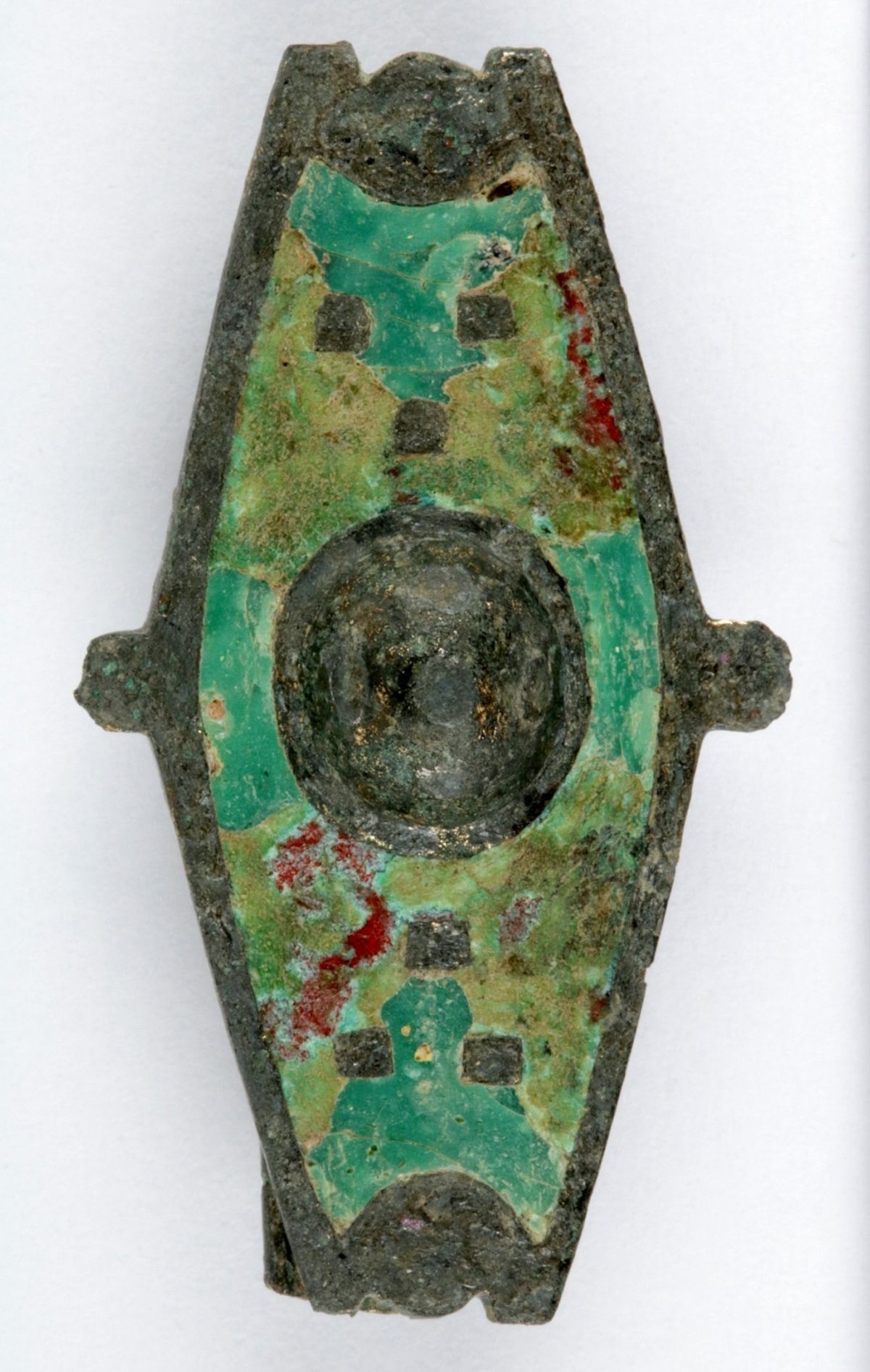 Rautenförmiges Metallobjekt mit grünlichen Einlagen aus Emaille. Das Metall ist korrodiert