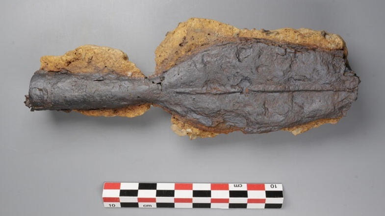 Längliches Objekt mit breitem, flachem Ende und schmalem Griff, auf grauem Untergrund liegend, darunter ein Maßstab mit schwarz-weißen und roten Feldern