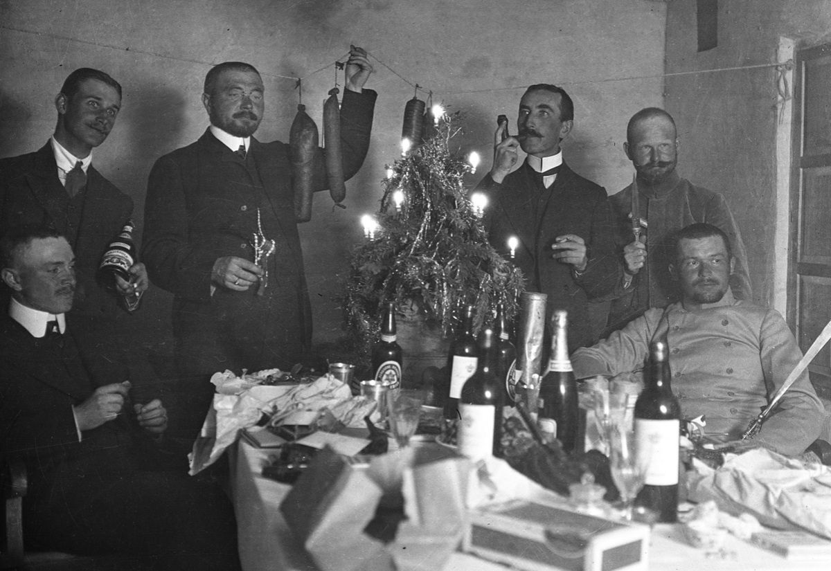 Mehrere Männer in Anzügen und Uniformen sitzen und stehen um einen Tisch mit Flaschen, Gläsern und einem kleinen Weihnachtsbaum mit Kerzen.