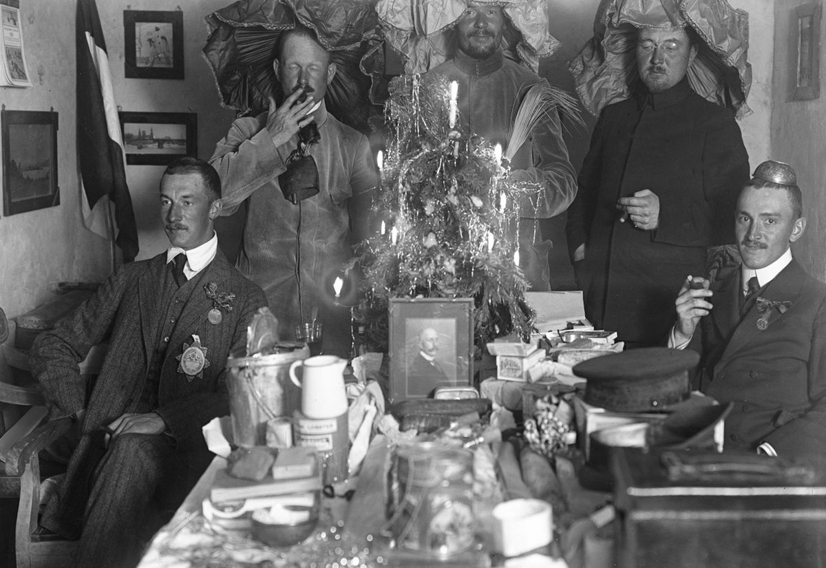 Mehrere Männer in Anzügen und Uniformen sitzen und stehen um einen Tisch mit einem kleinen geschmückten Weihnachtsbaum, Kerzen und verschiedenen Gegenständen.