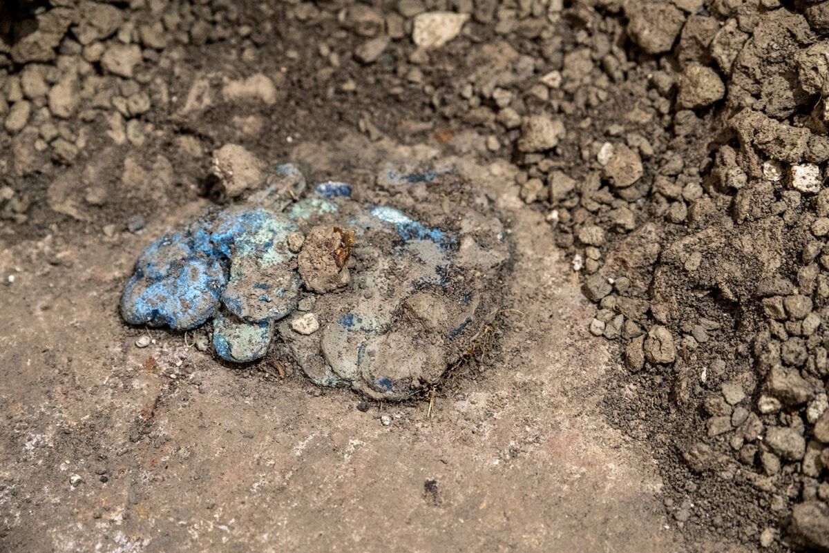 Im Mittelpunkt liegt ein Haufen antiker Münzen, die teilweise mit Erde bedeckt sind. Die Münzen zeigen eine bläuliche Patina, die auf Alter und Oxidation hinweist. Der Boden besteht aus festem, steinigem Material mit vielen kleinen, unregelmäßigen Steinen und Erdklumpen.