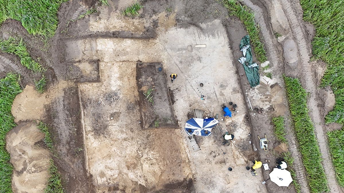 Luftaufnahme einer archäologischen Ausgrabungsstätte mit mehreren Personen, zwei Sonnenschirmen und umgebender Vegetation.