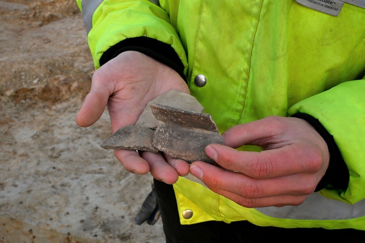 Hände einer Person in gelber Warnjacke halten mehrere Bruchstücke von graubrauner Keramik.