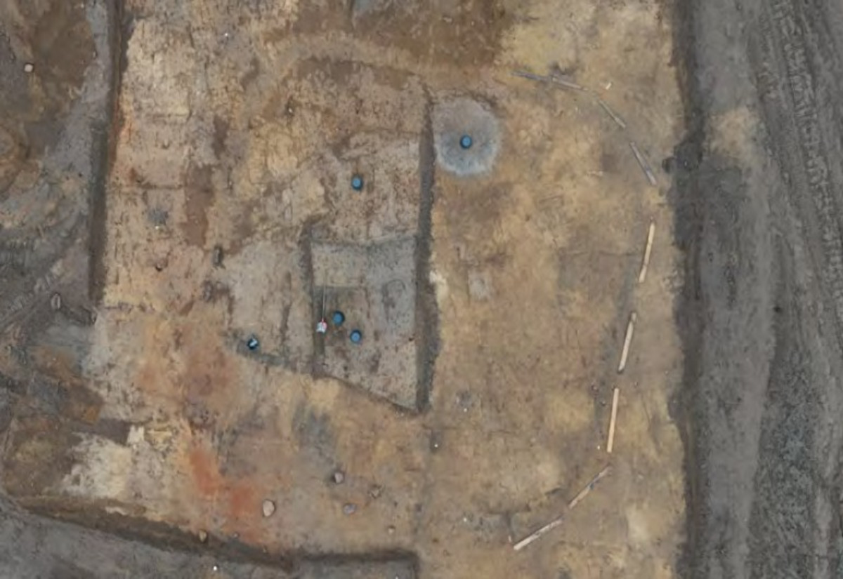 Luftaufnahme der archäologischen Ausgrabungsstätte Seddin mit einem rechteckigen Bereich in der Mitte, der durch dunklere Erde markiert ist. Mehrere blaue Markierungen sind innerhalb und um diesen Bereich verteilt. Rechts ist eine gebogene Linie aus hellen Stäben oder Markierungen zu sehen, die einen Teil des Geländes abgrenzt. Die Oberfläche zeigt verschiedene Farbtöne von Braun, Grau und Orange.