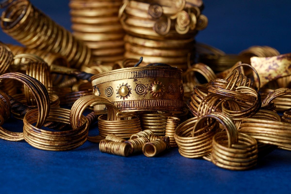 Mehrere goldene Armreifen und Spiralen liegen auf einer dunkelblauen Fläche. Im Vordergrund befindet sich ein breiter Armreif mit eingravierten Sonnenmotiven und umlaufendem Muster. Die Schmuckstücke bestehen aus glänzendem Metall und zeigen unterschiedliche Größen und Wicklungen.