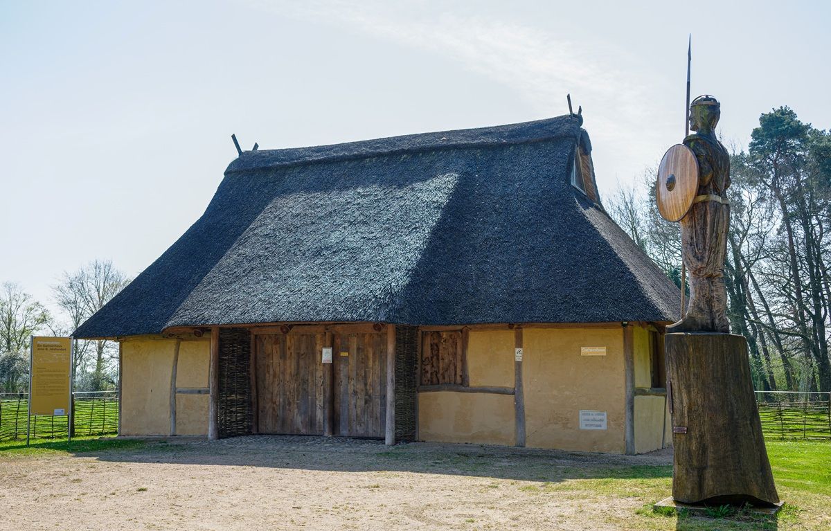 Im Vordergrund steht eine große Holzskulptur mit Schild und Speer auf einem Sockel. Dahinter befindet sich ein langgestrecktes Gebäude mit Reetdach, Holztüren und Lehmwänden. Das Gebäude ist von einem Zaun umgeben, und im Hintergrund sind Bäume und blauer Himmel sichtbar.