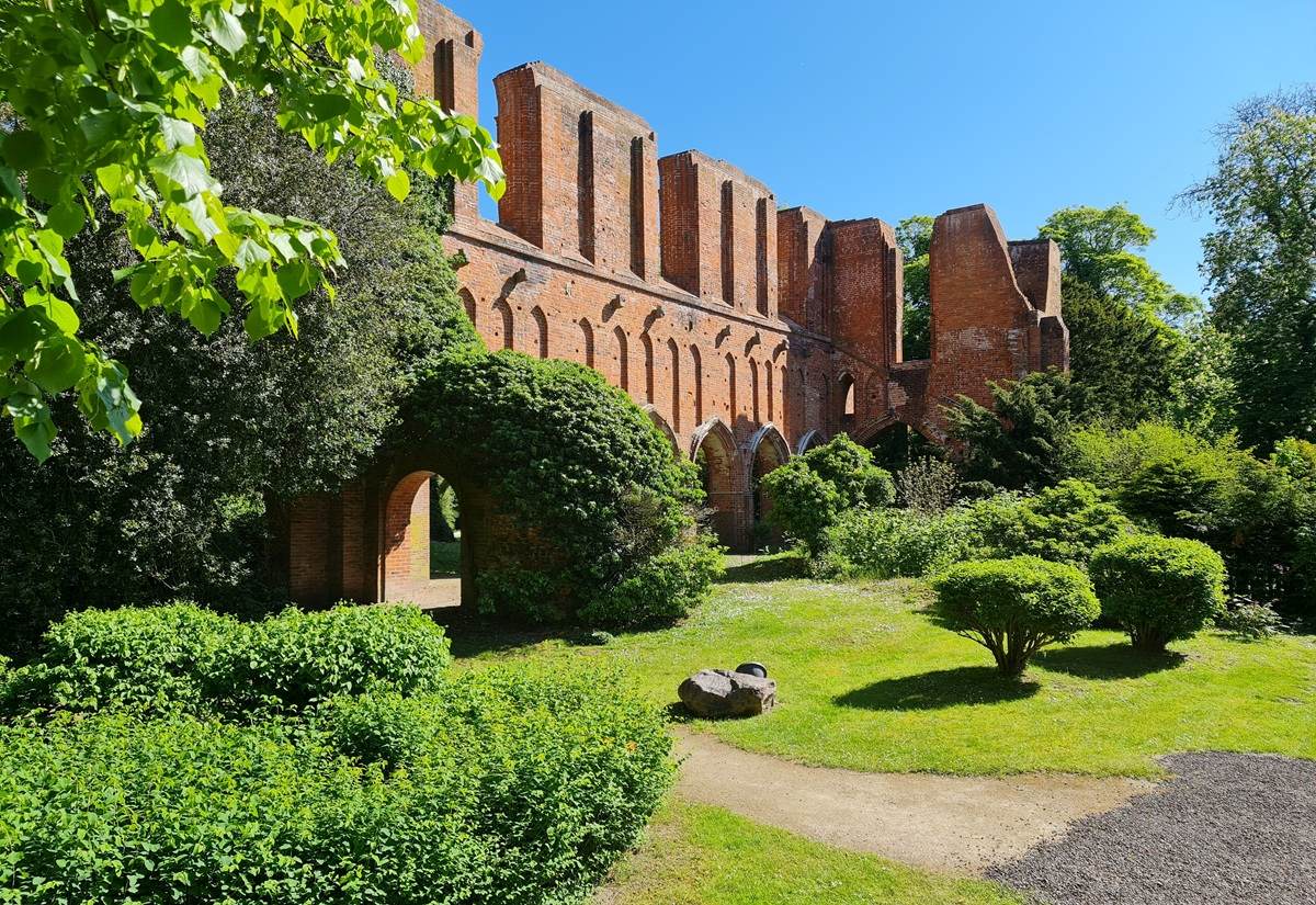 Rote Backsteinruine mit hohen, rechteckigen Fensteröffnungen steht in einer gepflegten Grünanlage. Dichte Büsche und Bäume rahmen das Bauwerk, Sonnenlicht fällt auf den Rasen und die Fassade. Ein Rundbogenportal ist im unteren Bereich sichtbar, daneben liegen zwei große Steine. Der Himmel ist klar und blau.