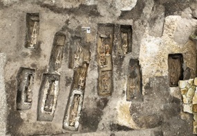 Mehrere rechteckige Grabkammern sind in einem Erdreich angeordnet. In den Kammern liegen menschliche Skelette, die vollständig erhalten wirken. Die Knochen sind hell und heben sich deutlich vom dunkleren Boden ab. Ein kleiner, quadratischer Maßstab liegt zwischen den Gräbern zur Orientierung. Rechts im Bild sind einige Steine zu sehen, die eine Mauer oder Begrenzung andeuten.