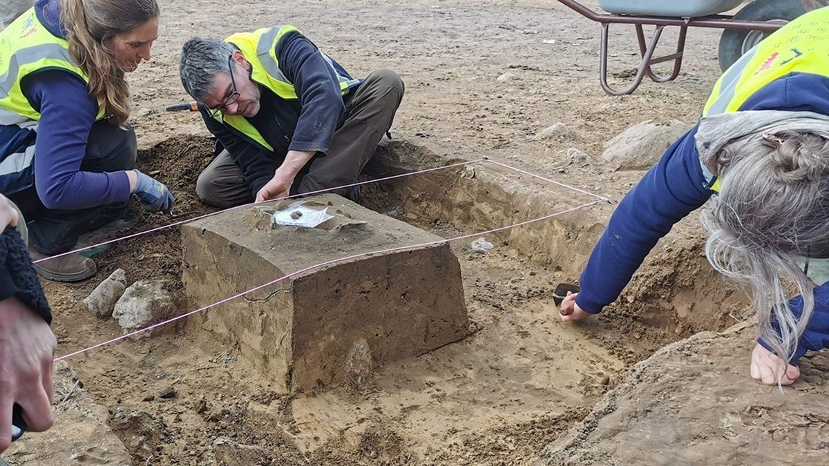 Drei Personen in Warnwesten arbeiten auf einer Ausgrabungsstätte. Sie knien auf dem Boden und untersuchen einen rechteckigen, teilweise freigelegten Erdblock, der von einer Schnur umrahmt ist. Auf dem Block liegt ein kleines, rundes Objekt. Im Hintergrund steht eine Schubkarre auf sandigem Boden.