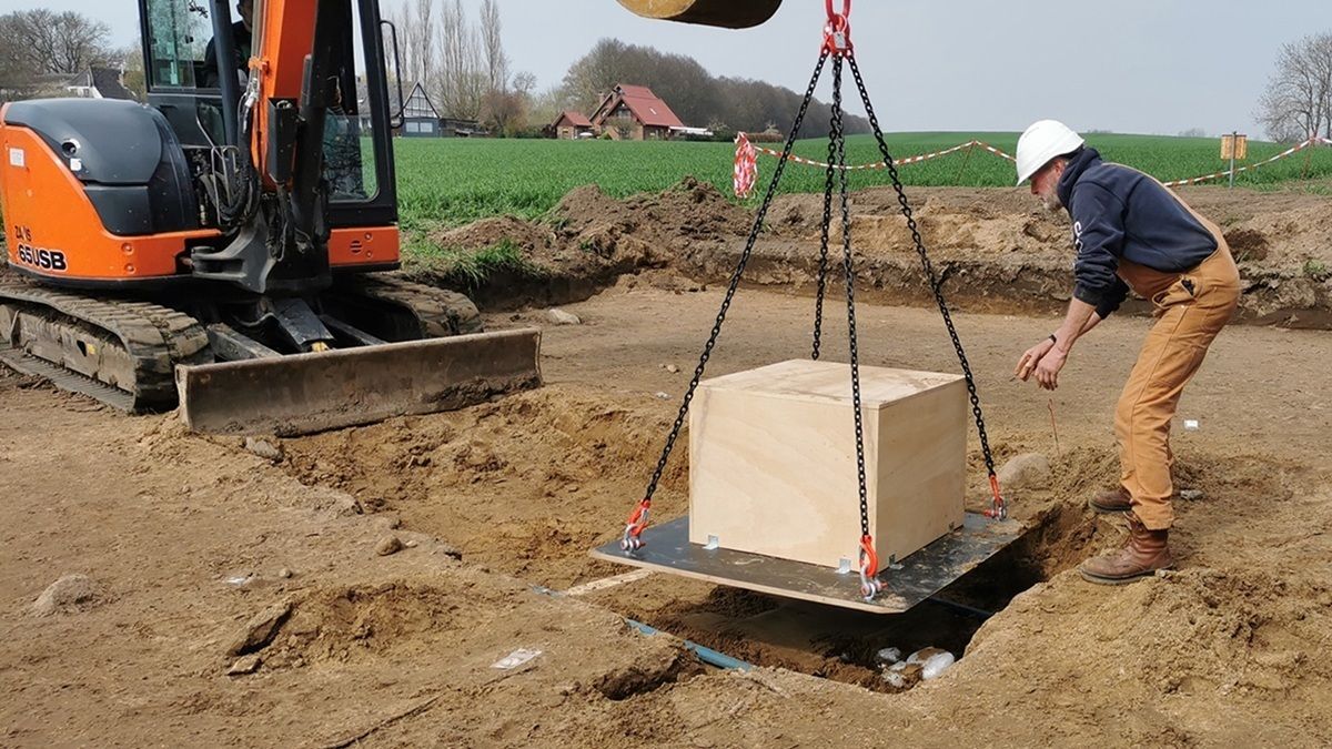 Ein großer Holzkasten wird mithilfe von Ketten und einem Bagger aus einer rechteckige Grube gehoben. Ein Mann mit weißem Schutzhelm und Arbeitskleidung steht daneben und beobachtet den Vorgang. Im Hintergrund sind Felder, einige Bäume und Gebäude zu sehen.
