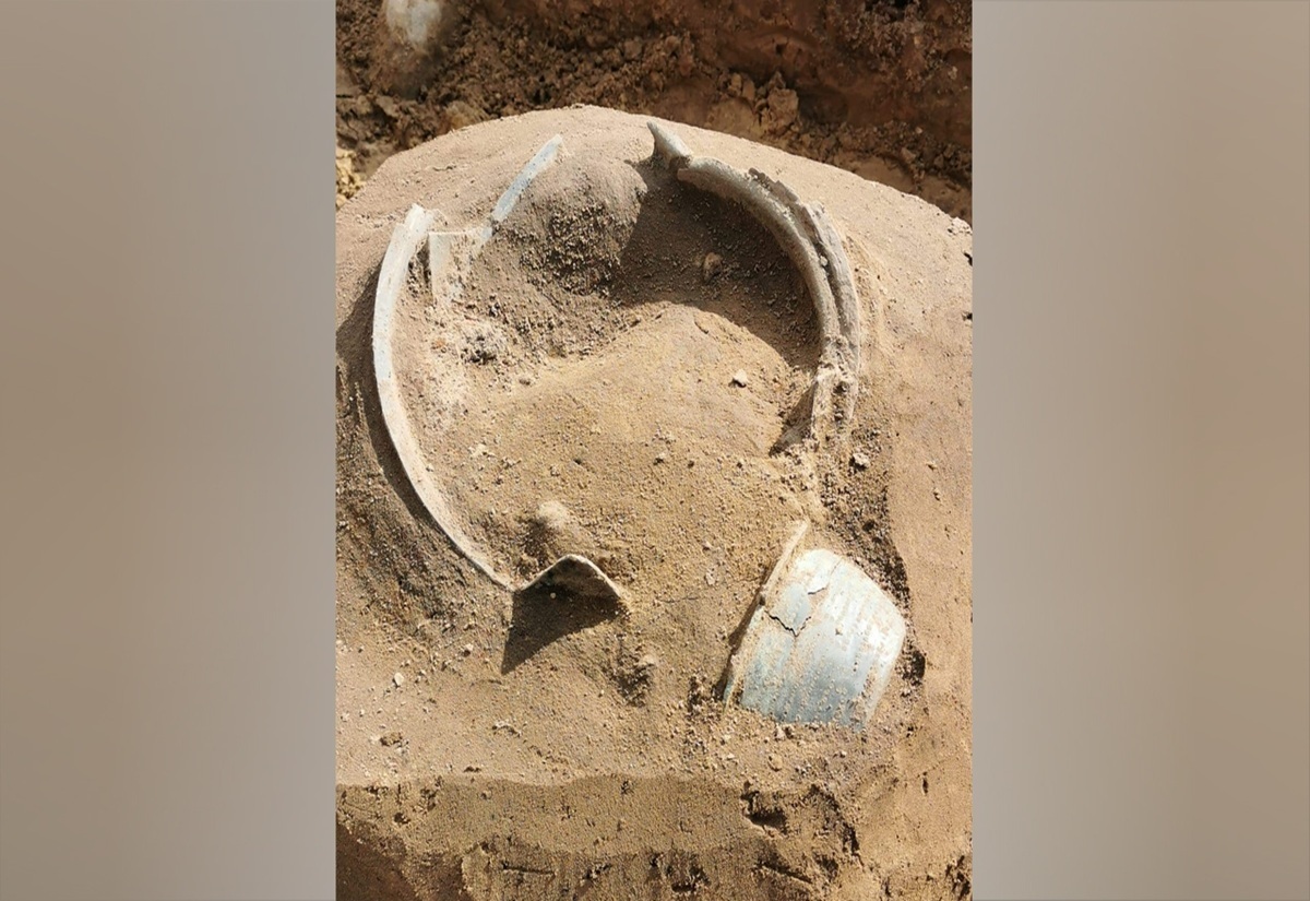 Ein bronzener Kessel liegt halb freigelegt in sandiger Erde. Der Kessel ist beschädigt, mehrere Bruchstücke sind sichtbar. Die Oberfläche zeigt eine grünlich-blaue Patina, die für Bronze typisch ist. Die Form des Gefäßes ist rund, mit einem breiten Rand und einer bauchigen Wandung. Der Kessel ist von losem Erdmaterial umgeben.