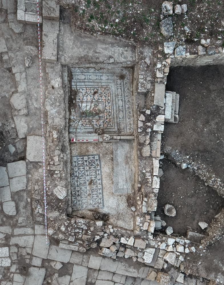 Luftaufnahme einer archäologischen Ausgrabungsstätte mit freigelegtem Mosaikboden. Das Mosaik zeigt geometrische Muster und farbige Ornamente in einem rechteckigen Feld. Um das Mosaik sind Steinplatten und Mauerreste sichtbar. Die Fundstelle ist von Erde und Steinen umgeben, ein rot-weißes Band markiert einen Bereich am Rand.