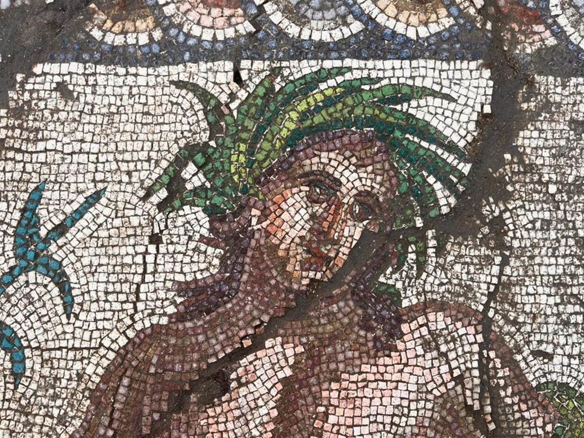 Mosaik mit einer menschlichen Figur, die von kleinen, unterschiedlich gefärbten Steinchen gebildet wird. Die Figur hat einen nach vorn geneigten Oberkörper, einen nachdenklichen Gesichtsausdruck und grüne, blattartige Elemente im Haar. Im oberen Bereich verläuft ein ornamentales Band mit runden Mustern.