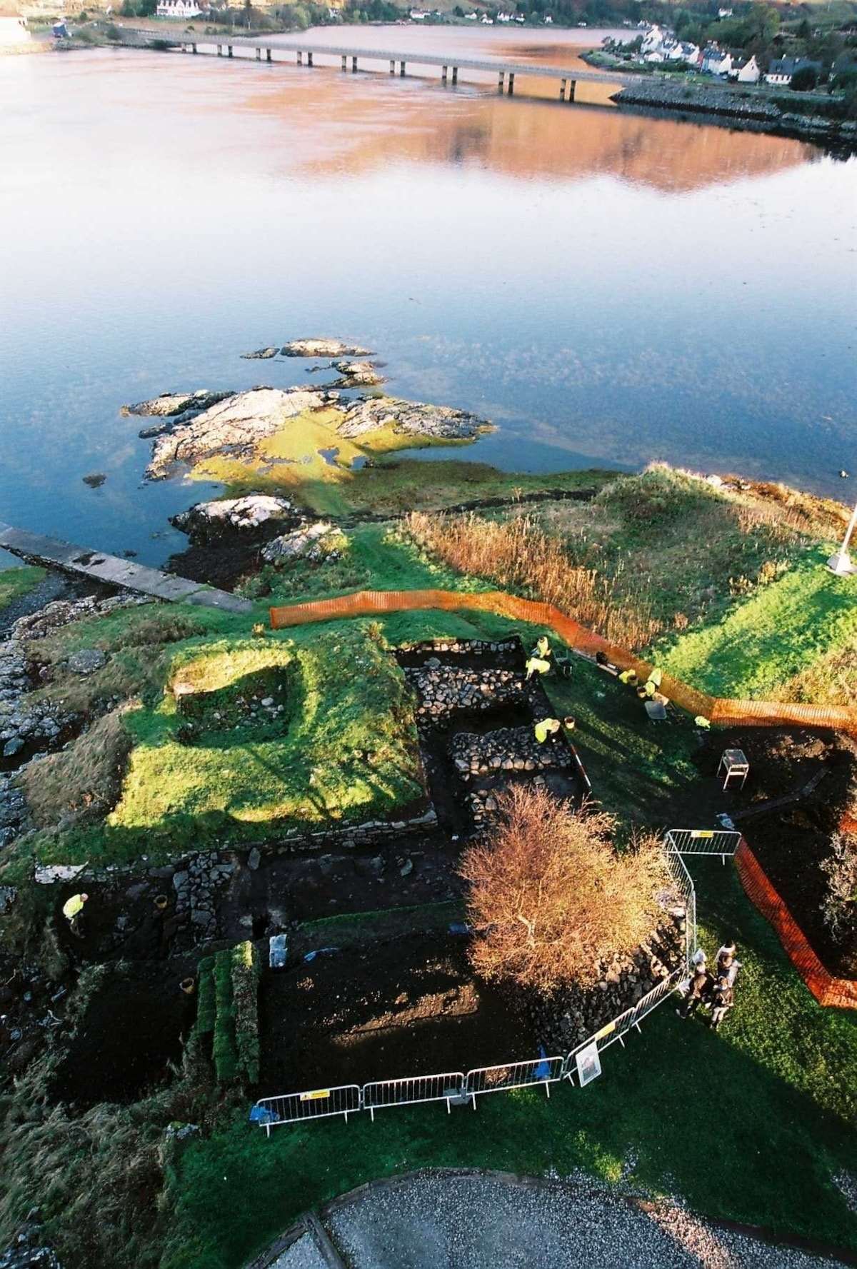 Luftaufnahme einer archäologischen Ausgrabungsstätte mit rechteckigen Gruben und Wegen am Ufer eines Flusses bei Sonnenlicht.