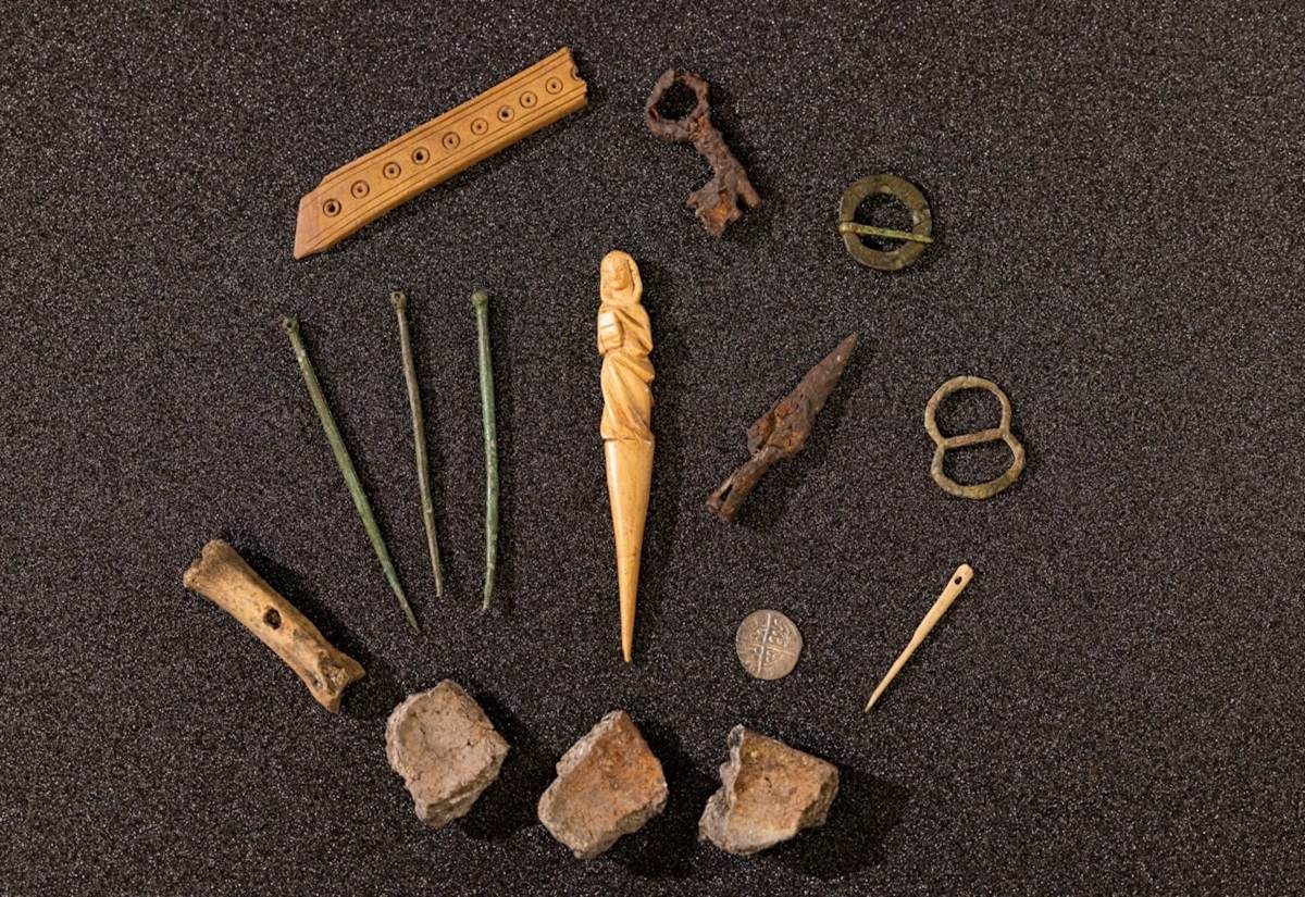 Verschiedene archäologische Fundstücke auf dunklem Untergrund, darunter eine geschnitzte Figur, Gewandnadeln, Schlüssel, Münze und Keramikfragmente.