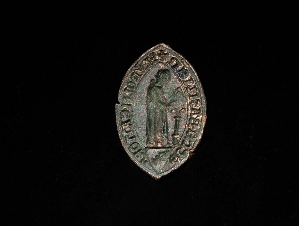 Ovales, bronzefarbenes Siegel mit einer stehenden Figur und umlaufender Inschrift auf schwarzem Hintergrund.