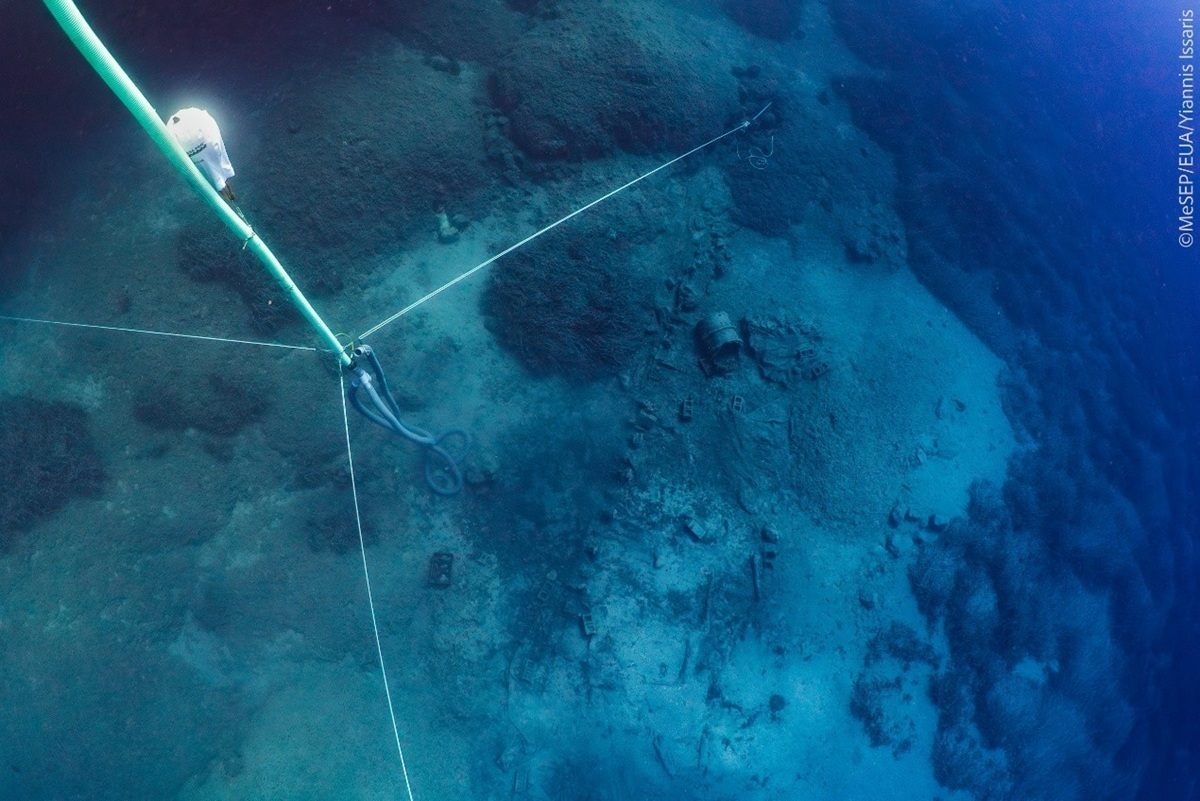 Unterwasseraufnahme eines Schiffswracks auf sandigem Meeresgrund. Mehrere gespannte Seile verlaufen diagonal über das Bild. Im Zentrum ist ein größeres, zylindrisches Objekt zu erkennen, das teilweise im Sand liegt. Die Umgebung ist von Felsen und Sedimenten geprägt, das Wasser wirkt klar und blau. In der linken oberen Ecke ist ein technisches Gerät an einem der Seile befestigt.