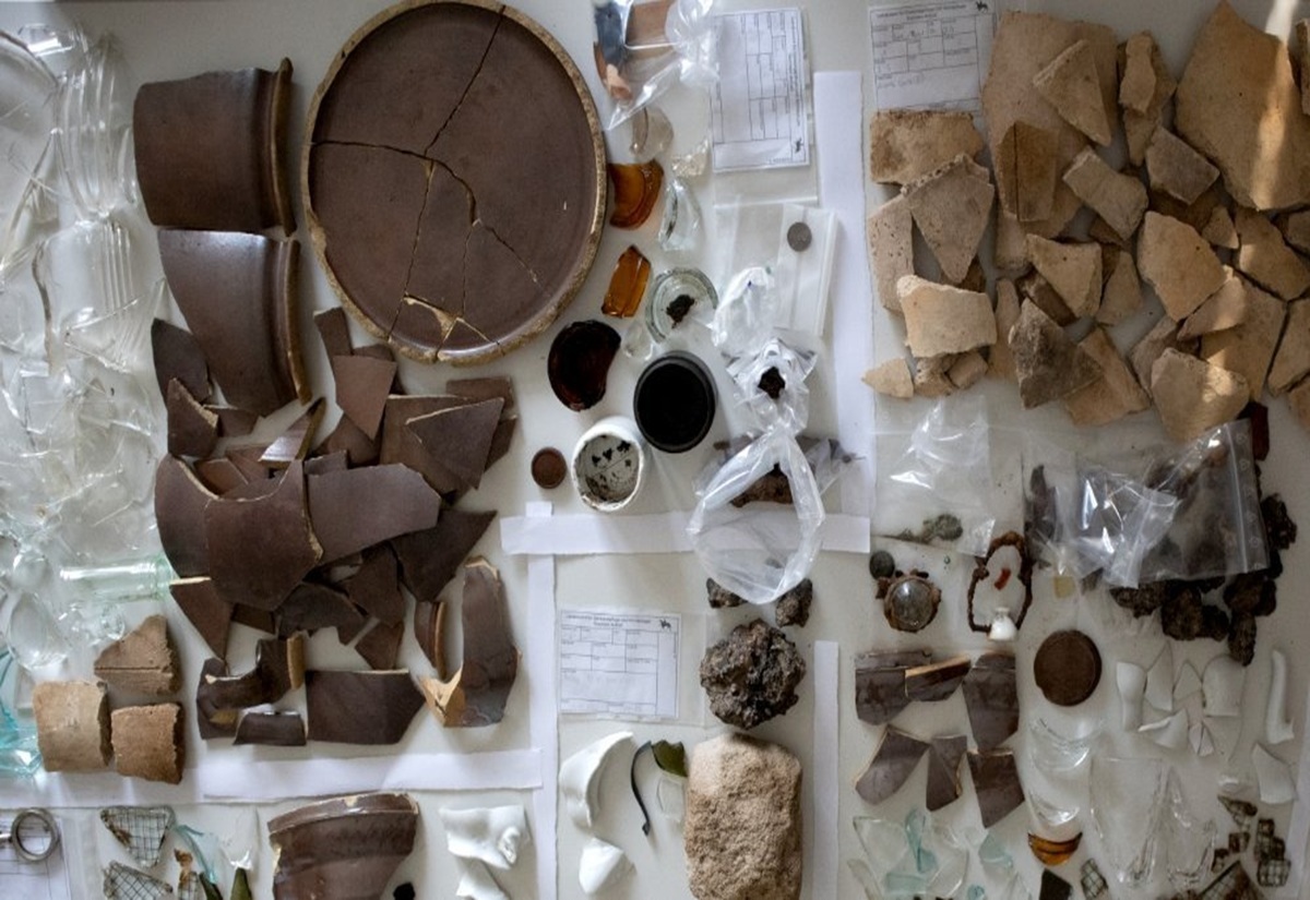 Auf einer weißen Fläche liegen zahlreiche archäologische Fundstücke, darunter zerbrochene Keramik- und Porzellanscherben, Glasfragmente, kleine Metallobjekte und mehrere beschriftete Fundzettel. Einige Funde sind in durchsichtigen Kunststoffbeuteln verpackt. Die Objekte sind in Gruppen sortiert und nehmen die gesamte Bildfläche ein.