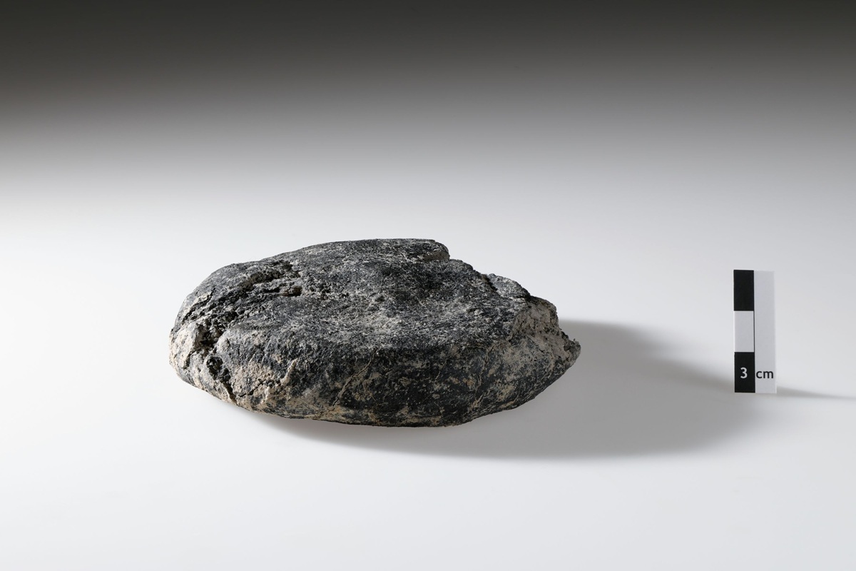 Ein rundes, flach gedrücktes, dunkel verfärbtes Objekt mit rauer Oberfläche liegt auf einer hellen, glatten Fläche. Rechts daneben steht ein Maßstab mit abwechselnd hellen und dunklen Segmenten sowie einer Markierung für drei Zentimeter. Das Objekt wirkt verkohlt und erinnert in Form und Struktur an ein Brotlaib.