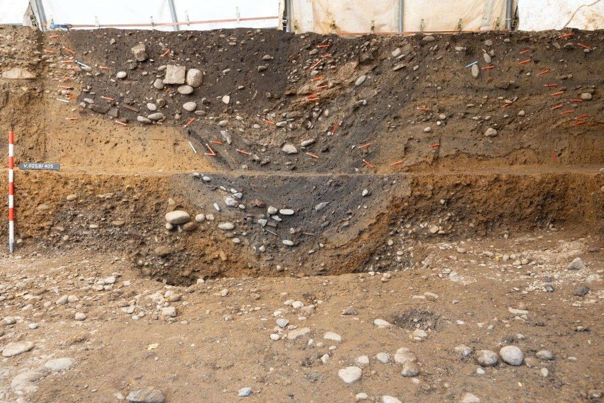 Querschnitt einer archäologischen Grabungsstelle mit deutlich sichtbaren Erdschichten. Im oberen Bereich sind dunkle, fast schwarze Erdschichten mit eingelagerten Steinen und vereinzelten rötlichen Markierungen zu erkennen. Darunter folgt eine hellere, gelblich-braune Schicht, die ebenfalls Steine enthält. Im Vordergrund liegt ein unebener, steiniger Boden. Links steht ein rot-weiß gestreifter Messstab, daneben ein kleines Schild mit weißer Schrift auf blauem Grund. Im Hintergrund ist eine helle Abdeckung gespannt.