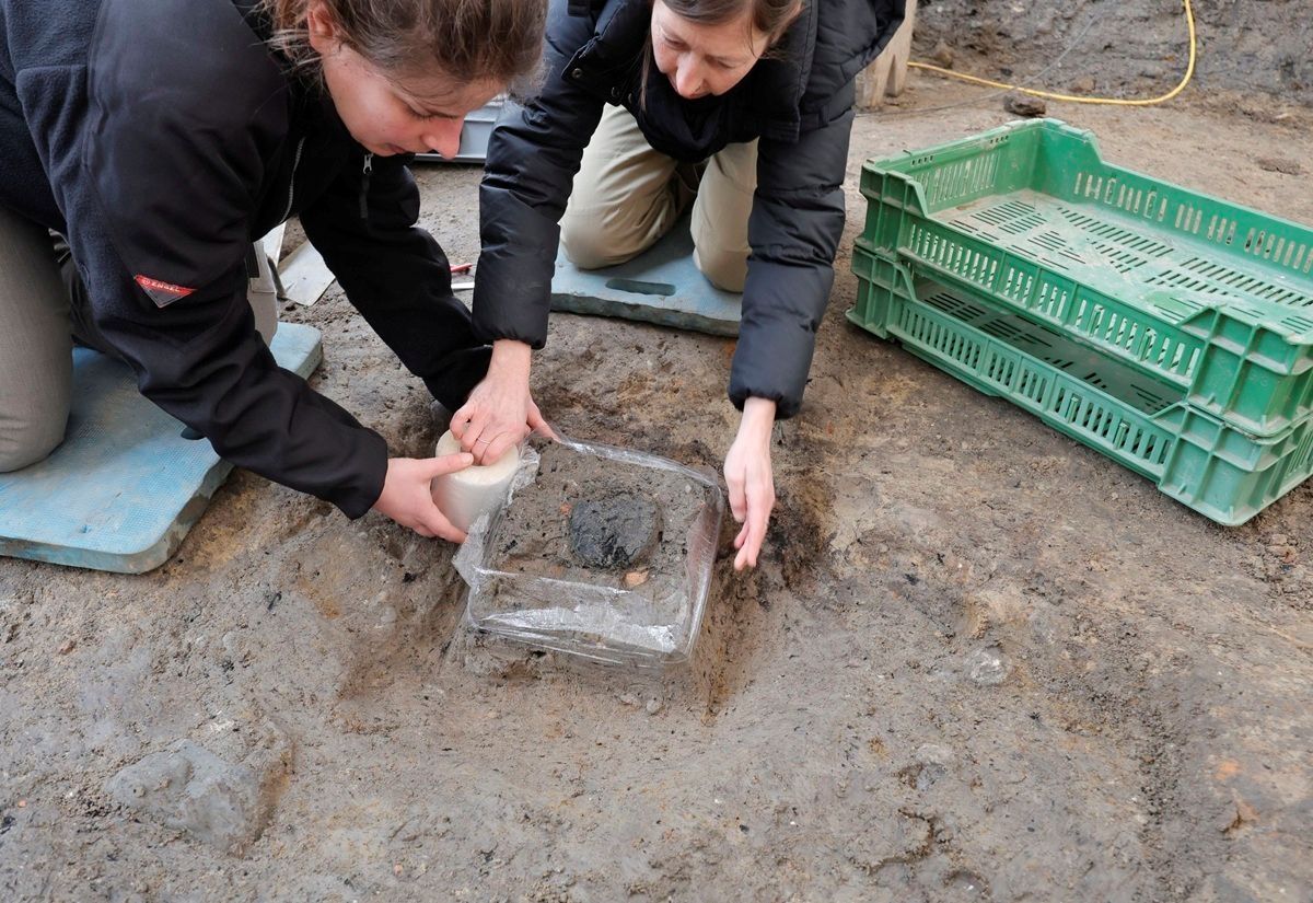 Zwei Personen knien auf dem Boden einer Ausgrabungsstätte. Sie tragen dunkle Jacken und helle Hosen. In einer rechteckigen, flachen Grube liegt ein durchsichtiger Kunststoffrahmen, in dem sich ein dunkler, runder Gegenstand befindet. Eine Person hält einen kleinen Pinsel, mit dem sie vorsichtig an dem Gegenstand arbeitet. Im Hintergrund stehen grüne Kunststoffkisten übereinandergestapelt. Der Boden ist erdig und feucht.