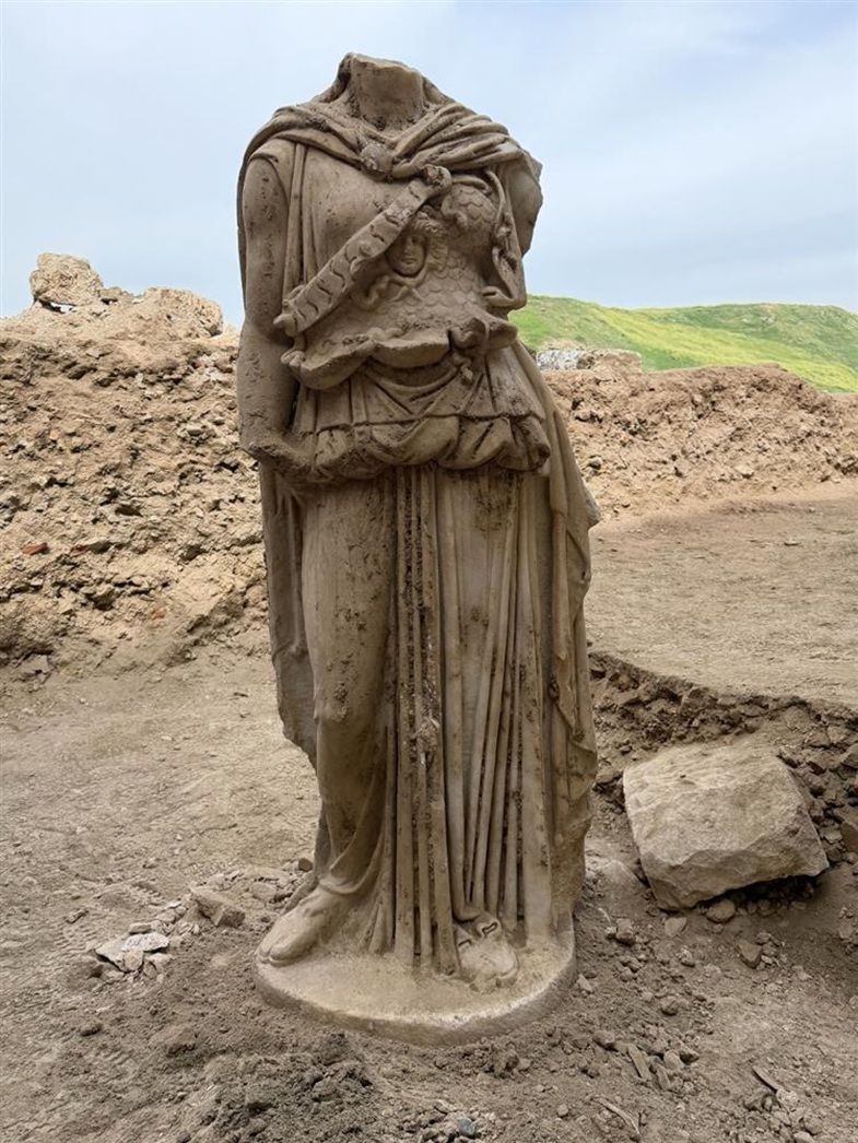 Eine antike Statue ohne Kopf steht aufrecht auf staubigem Boden vor einem Hintergrund aus Erdaufschüttungen und grünem Hügel. Die Figur trägt ein langes, gefälteltes Gewand und einen Umhang, der über die linke Schulter gelegt ist. Die Oberfläche der Statue ist stellenweise beschädigt und verschmutzt.