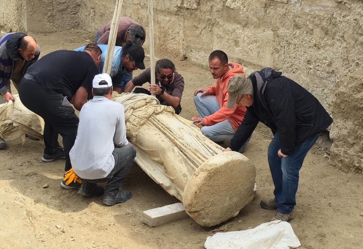 Eine antike Statue liegt auf dem Boden einer Ausgrabungsstätte. Mehrere Personen beugen sich über die Statue und arbeiten gemeinsam daran, sie zu bergen. Die Statue ist aus hellem Stein gefertigt und zeigt eine aufwendig gestaltete Figur mit langen Gewändern. Der Hintergrund besteht aus einer sandfarbenen, steinernen Wand.