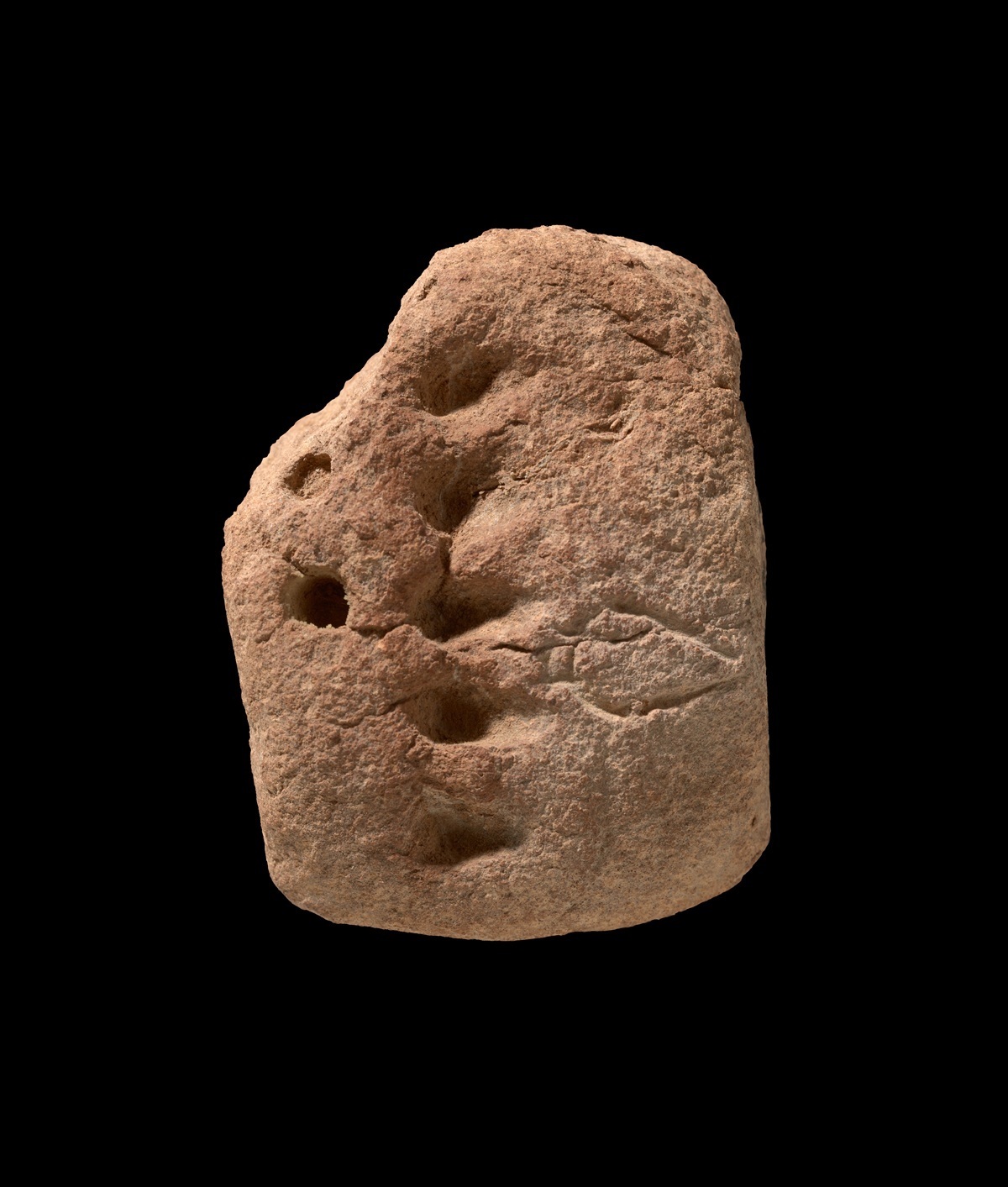 Unregelmäßig geformter, aufrecht stehender Stein mit mehreren runden Vertiefungen und einer eingeritzten, länglichen Linie mit Querverbindungen auf der Vorderseite. Die Oberfläche wirkt rau und porös. Der Hintergrund ist einfarbig schwarz.