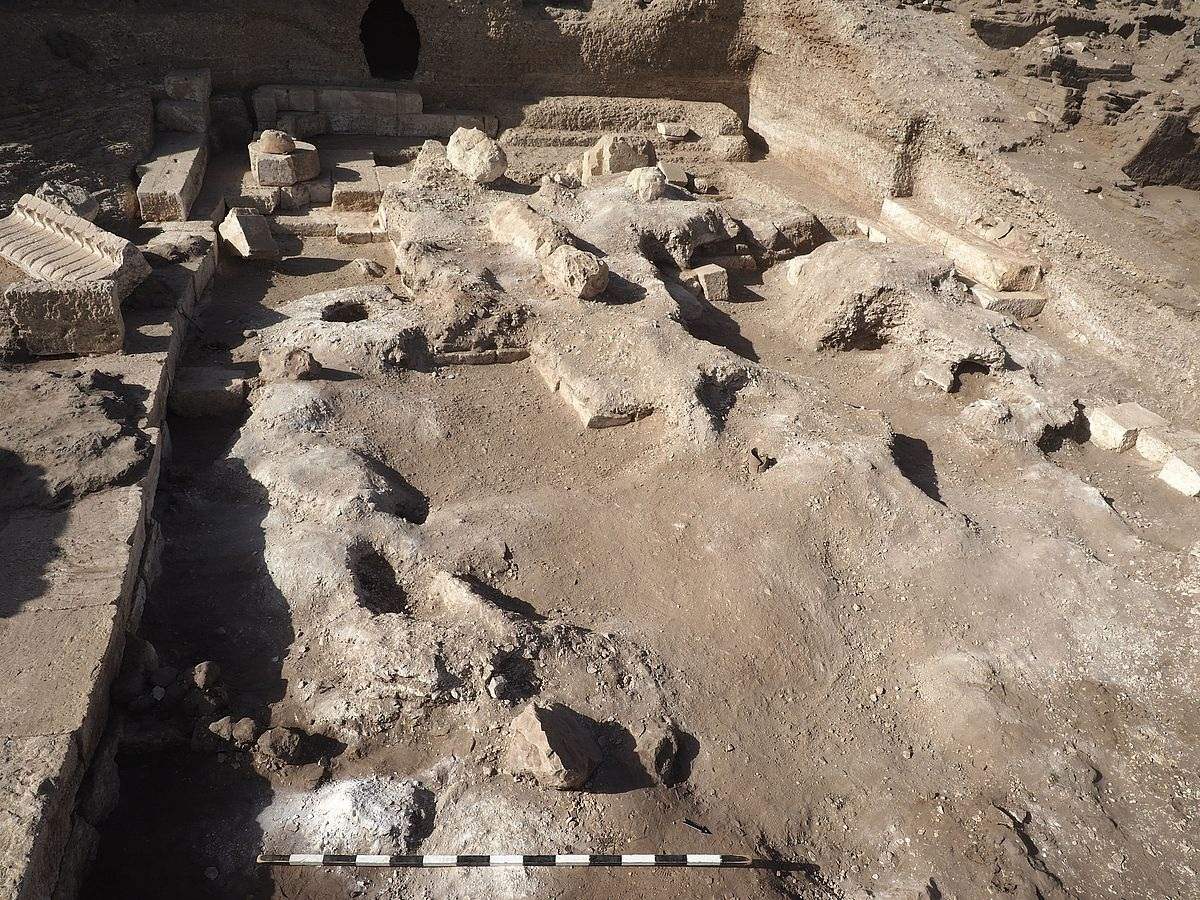 Archäologische Ausgrabungsstätte mit freigelegten Fundamenten und Mauerresten aus hellem Stein. Die Fläche ist uneben und von Erdschichten sowie einzelnen Steinen und Blöcken durchzogen. Im Hintergrund sind weitere Mauern und eine dunkle Öffnung zu sehen. Im Vordergrund liegt eine Maßskala.