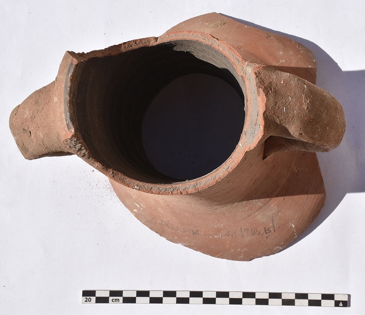 Fragment eines Tonkrugs mit zwei seitlichen Henkeln, von oben fotografiert. Die Oberfläche zeigt eine matte, rötlich-braune Färbung und leichte Gebrauchsspuren. Am unteren Rand sind feine, eingeritzte Zeichen erkennbar. Im Vordergrund liegt eine Maßskala mit schwarz-weißen Feldern und Zentimeterangaben.