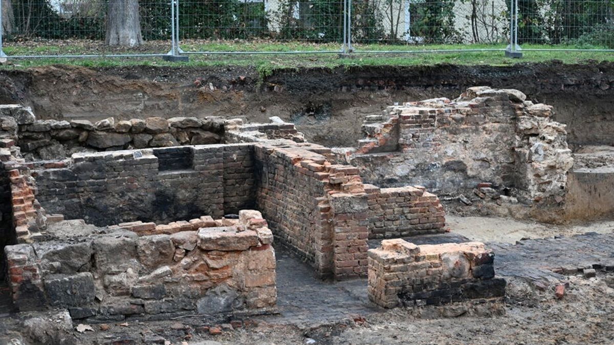 Freigelegte, teilweise erhaltene Mauerreste aus Backstein und Naturstein in einem archäologischen Ausgrabungsbereich. Die Mauern bilden rechteckige und unregelmäßige Strukturen, die unterschiedliche Höhen aufweisen. Im Hintergrund ist ein Metallzaun zu sehen, der das Areal abgrenzt. Im oberen Bereich des Bildes sind Gras und Baumstämme sichtbar.