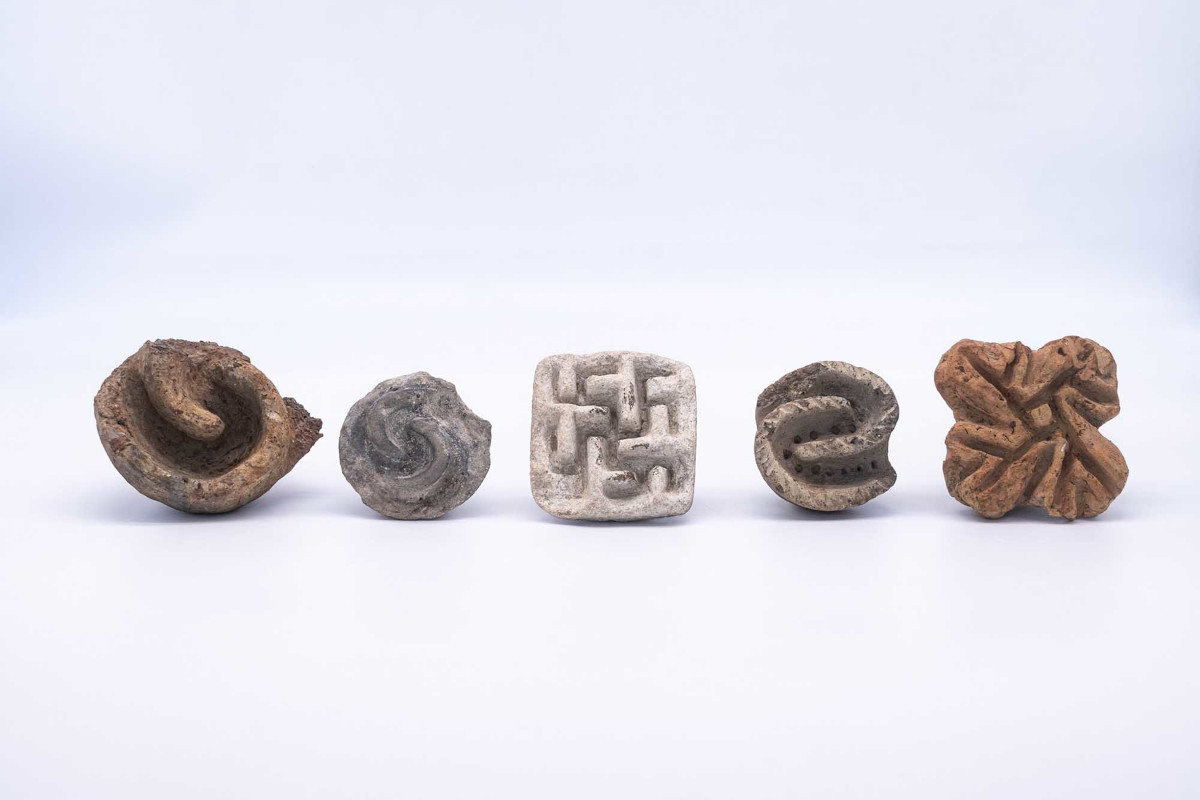 Fünf kleine Objekte Terracotta liegen nebeneinander auf einer weißen Fläche. Jedes Objekt zeigt eine andere, deutlich erkennbare Reliefstruktur mit verschlungenen, labyrinthartigen Mustern. Die Formen variieren von rund bis eckig, die Oberflächen sind teils glatt, teils rau. Die Farbpalette reicht von hellgrau bis ockerbraun.