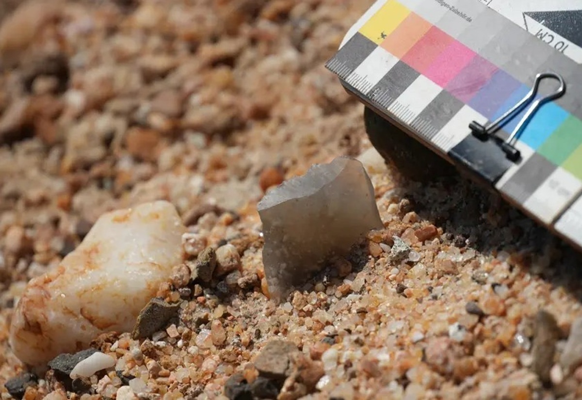 Kleines, spitz zulaufendes, graues Steinfragment liegt auf sandigem, kiesigem Untergrund neben einem größeren weißen Stein und einem Farb- und Grauwertmaßstab mit Klammer.